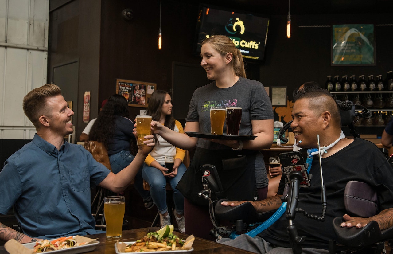 a waitress serves a beer to a man in a bar seated with his friend who is in a wheelchair with assistive devices